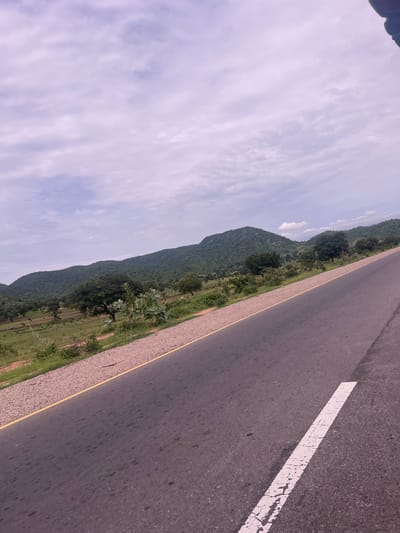 This media file depicts a roadside view in Minda, Nigeria. The scene is dominated by a dark asphalt road running diagonally from the lower right to the upper left corner of the frame. A broken white line, characteristic of road markings, is visible on the right side of the road. A yellow line also demarcates the edge of the road. To the left of the road, a gravel shoulder transitions into a lush green landscape. Rolling hills covered in dense vegetation rise from the green fields. The sky above is filled with light, wispy clouds, suggesting a bright day, possibly late morning or early afternoon. The overall impression is one of a rural or semi-rural setting, highlighting the road as a passage through a natural, verdant environment. There is no visible text in the image. The perspective suggests the image was taken from a moving vehicle.
