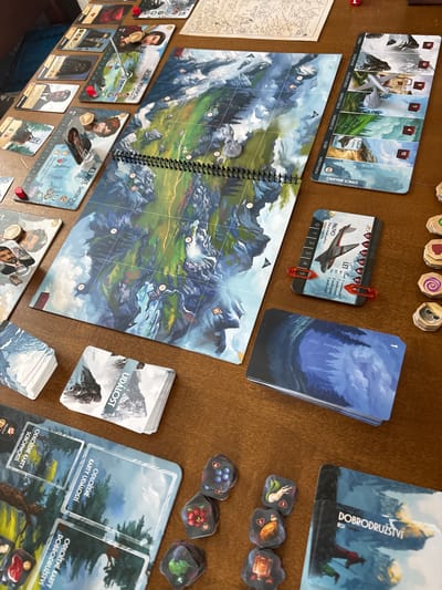 This is a top-down, close-up photograph of a board game spread across a wooden surface. The game features a large, spiraling board depicting a mountainous, snowy landscape with various locations marked with numbers and symbols. Several decks of cards are visible, some fanned out, displaying artwork of forests, mountains, and characters. One card, secured with a red clip, shows a vintage airplane and text in Czech, indicating "FUEL" and details about potential consequences. Another card, with the title "UDÁLOST" (Event), shows a desolate mountain scene.
Game pieces, including small wooden tokens and miniatures representing airplanes and possibly figures, are scattered on the board. Individual character cards are also laid out, featuring portraits of people with names like "JESSIE WILDEROVÁ" and "CLAIRE SMITHOVÁ," along with their respective locations, "SAN FRANCISCO" and "SEATTLE." Smaller tokens, some square and some hexagonal, depict various items such as fruits and vegetables. A card with the title "DOBRODRUŽSTVÍ" (Adventure) shows a traveler with a dog.
The scene suggests a tabletop gaming session in progress, likely in a casual, indoor setting given the lighting and wooden table. The artwork suggests a theme of exploration and adventure, possibly in a wilderness or survival context. The presence of Czech text indicates the game is likely a localized version.