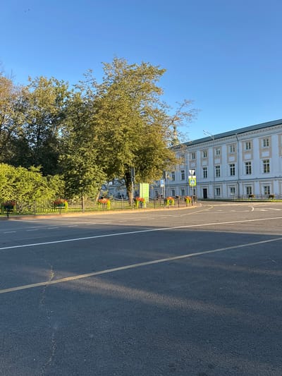 The image depicts a wide asphalt road in Yazvitsevo, Russia, with clear markings indicating lanes and a yellow center line. The road curves to the right, leading towards a large, white building with a green roof and numerous windows. The building appears to be a historical structure, possibly a museum or an educational institution, judging by the signboards and a pedestrian crossing signal visible near it.
A row of decorative planters with red flowers lines a black metal fence that separates the road from a green area. Behind the fence, mature trees with lush green foliage dominate the landscape, with some branches extending over the road. The sky is a clear, bright blue, suggesting a sunny day. The lighting indicates it's likely morning or late afternoon, casting long shadows across the road. No people are clearly visible, but distant figures might be present near the building. The overall scene is serene and urban, with a blend of natural elements and man-made structures.