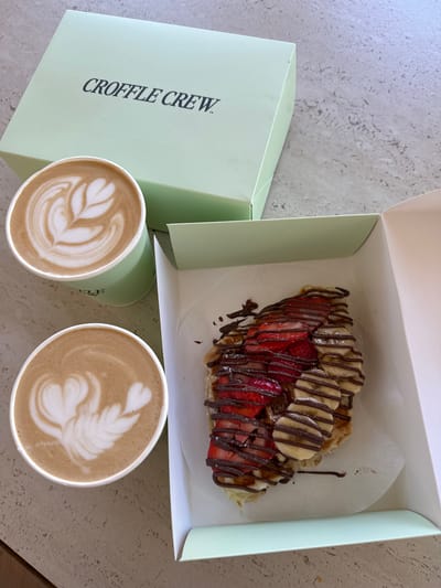 In Happy Valley, Oregon, United States, a light green box with the inscription "CROFFLE CREW" is placed on a speckled surface. Next to it are two cups of coffee with latte art. The coffee on top features a leaf-shaped design, while the one below has a swan-like design. In an open light green and white box, a croffle is presented, topped with sliced strawberries and bananas, and drizzled with chocolate.