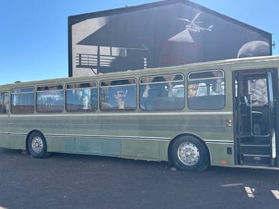 The scene depicts a static outdoor arrangement at a film studio in Ouarzazate, Morocco, under a clear blue daytime sky. An olive-green vintage-style passenger bus is parked on dark, unpaved ground. Its front passenger door is open, revealing interior steps and a large, monochrome human face painted on the bus exterior near the door. The bus windows display posters for the television series 'Prison Break,' including character images and the show's title, with light-colored curtains visible behind some panes. The right rear tire of the bus appears deflated and rests on a dark block. In the background, a large, dark-colored building with a pitched roof features a mural. This mural depicts a white helicopter with a searchlight beam, the words 'PRISON BREAK' in red capital letters, and the silhouette of a building with a balcony. A large, dark, rounded object is partially visible on the far right side of the building. No individuals are present or engaged in any actions or interactions.