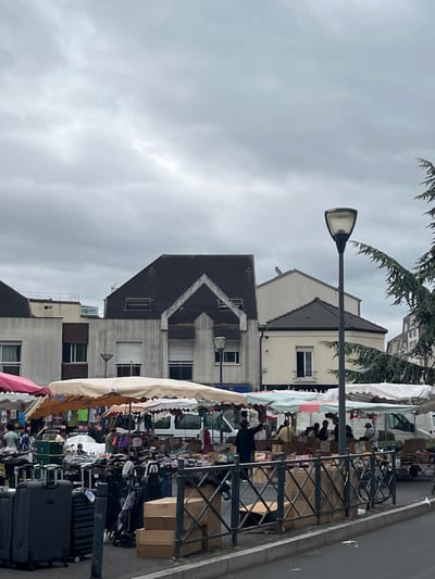 This image depicts an outdoor market scene in the city of Villeparisis, France, under an overcast sky. The foreground is dominated by a paved area with a low metal fence, behind which are several large, dark suitcases and some cardboard boxes. Beyond the fence, a row of market stalls with colorful awnings is set up. Various goods are displayed on tables and racks, including clothing. People are visible at the stalls, some interacting with each other and browsing. Behind the market stalls, there are buildings, including a multi-story building with a distinctive dark, gabled roof and several windows. A lamppost stands in the middle ground, and trees with dark green foliage are visible on the right side of the image. The overall atmosphere appears to be that of a typical, bustling market day.