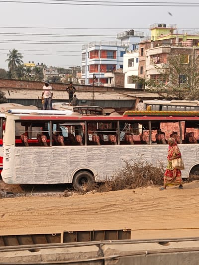 The scene in Boliarpur, Bangladesh, depicts an urban environment under a clear sky. In the foreground, a sandy embankment with visible tire tracks slopes downward, partially obscuring concrete barriers and metal structures at the bottom. A woman, dressed in a red and yellow sari with blue sandals, walks from left to right on this embankment, carrying a light-colored bag over her left shoulder.
In the midground, a white bus with a prominent red stripe is stationary on a dirt patch. Its exterior displays a textured, light grey finish, and rows of patterned brown seats are visible through its open windows. Dry shrubbery is situated to the bus's right. Multiple overhead utility lines crisscross the frame above the midground.
Behind the bus, several individuals are positioned on elevated structures. One man, wearing a light-colored shirt and a hat, stands upright on a roof-level structure, holding an indistinct object. Another man in a dark t-shirt and light pants stands further back on an adjacent roof. Two additional men are seated on different roof structures further to the right, one wearing a patterned shirt.
The background features several multi-story buildings of varying colors, including light blue, yellow, and white, some exhibiting balconies and rooftop water tanks. A single palm tree is visible on the far left background, amidst other sparse trees and vegetation.