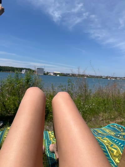 The image shows a first-person perspective of a person relaxing on a towel near a body of water. The foreground features the person's legs, which are tanned, and a colorful towel with a blue and green geometric pattern. The midground consists of green grass and vegetation, with a hint of wildflowers. In the background, a body of blue water stretches across the frame, with a distant shoreline featuring buildings, trees, and construction cranes. The sky above is a clear blue with scattered white clouds. The location appears to be a recreational area by the water, possibly within or near the city of Molodezhny, Russia, given the context of the prompt. The image suggests a sunny day, suitable for outdoor relaxation.
