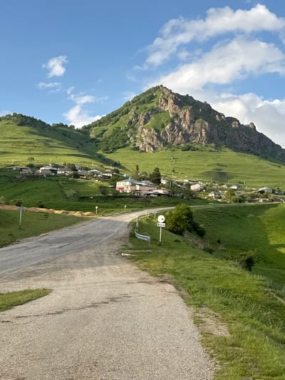 The image depicts a scenic view of the village of Nizhnyaya Mara, Russia, nestled amidst rolling green hills and dominated by a rugged, rocky mountain in the background. A paved road curves from the foreground into the village, which consists of scattered houses with light-colored roofs. The landscape is lush with vibrant green grass, suggesting a warm season. The sky above is a clear blue with scattered white clouds. A white road sign with black symbols is visible on the side of the road. The overall scene conveys a peaceful rural atmosphere.