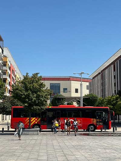 The image shows a street scene in Nîmes, France, on a sunny day. A red bus with yellow accents is parked on the right side of the frame. Several people are standing and walking near the bus. One person is seated on a low concrete wall in front of the bus, looking down at their phone. Another group of three people are standing near the bus entrance, with one holding a yellow bag.

Behind the bus and people, there are buildings. The building directly behind the bus appears to be a commercial establishment, with "CAPCINEMA" visible in large letters on its facade. To the left of this building, and further back, are other buildings with architectural features that suggest a European urban environment. One building has a distinct red and yellow patterned facade. Trees are scattered throughout the scene, adding greenery to the urban landscape.

The sky is clear and blue, indicating good weather. The sun is casting shadows, suggesting it is likely daytime, possibly mid-morning or afternoon. The overall atmosphere appears to be casual and everyday, with people going about their activities. The image is oriented vertically, making the bus appear sideways.