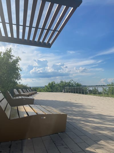 The image depicts an outdoor seating area with several lounge-style benches arranged in a row. The benches are made of wood and metal and are positioned on a paved patio. Beyond the patio, there is a railing overlooking a body of water, likely a river or lake, with trees and vegetation on the far bank. The sky is blue with scattered white clouds. In the distance, a person can be seen standing near the railing. The scene is bathed in sunlight, casting shadows from a wooden pergola overhead onto the patio and benches. The location is identified as the Shiryaevo village in Russia, specifically the Shiryaevo village museum-reserve.
