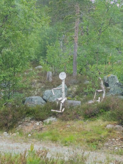 The image depicts a rustic, handcrafted figure made of birch branches, resembling a person, standing in a natural setting. The figure has a round, white head with a smiley face drawn on it. Its body is constructed from logs and branches, with arms outstretched. The figure is positioned amidst a rocky, wooded area with lush green foliage and scattered grey rocks.

The scene appears to be outdoors, likely in a forest or mountainous region, given the presence of trees, undergrowth, and rocks. The lighting suggests it could be daytime, possibly overcast, as there are no strong shadows. The overall atmosphere is serene and somewhat whimsical due to the presence of the handmade figure.

The figure itself is the central object of interest. Its construction suggests a folk art or decorative piece placed in the landscape. The surrounding environment is typical of a northern European forest, with birch trees and other greenery. The ground is covered with a mix of rocks, grass, and low-lying shrubs.

There are no people actively engaged in an activity, with the focus being solely on the static figure and its surroundings. The image captures a moment of stillness, highlighting the contrast between the man-made figure and the natural environment. No visible text is present in the image. The location context provided, Hovden, Norway, aligns with the visual cues of a northern, possibly Scandinavian, landscape.