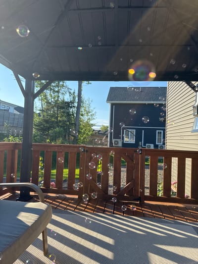 The image depicts a backyard deck with a gazebo. Numerous soap bubbles are floating in the air, some catching the sunlight and displaying iridescent colors. The deck is made of wooden planks and has a light-colored outdoor rug on one section. A gray outdoor chair is partially visible on the left side of the image. Beyond the wooden railing of the deck, a green lawn and trees are visible. Two houses are situated in the background, one dark blue and the other light-colored. The sky is clear and blue. The scene appears to be set in Dieppe, Canada, as indicated by the context of previous images. There are no people present in the image. The overall atmosphere is serene and suggests a warm, sunny day.