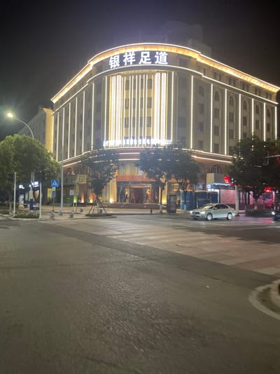 The image depicts a night scene at a street intersection in Dali City, China, focusing on a brightly illuminated multi-story building. The building, identified by its prominent rooftop sign as "银祥足道" (Yín Xiáng Zú Dào), features a light-colored facade with extensive vertical and horizontal strip lighting, accentuating its rounded corner and classical architectural elements such as columns. A large, well-lit entrance with steps is visible at its base.

The street in the foreground is asphalt, marked with pedestrian crosswalk lines. A light-colored sedan is positioned on the street, facing left. Several small trees are planted in a central median and along the sidewalks. Streetlights, a blue pedestrian crossing sign, and other street furniture are present. The scene indicates a commercial area. No individuals are clearly discernible in the immediate foreground or on the street.
