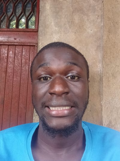 The image is a close-up, waist-up, outdoor selfie of a dark-skinned man with a short beard and dark hair. He is wearing a bright blue t-shirt and has his mouth open, revealing his teeth. He is looking directly at the camera with his eyes open. The background consists of a reddish-brown, textured wall on the right and a reddish-brown wooden door with a metal grate on the left. The lighting is natural and appears to be daylight, casting subtle shadows on the man's face and the wall. There is no specific indication of the city of Mbar, Nigeria, in the image itself, nor any other urban or rural indicators that could place it there.