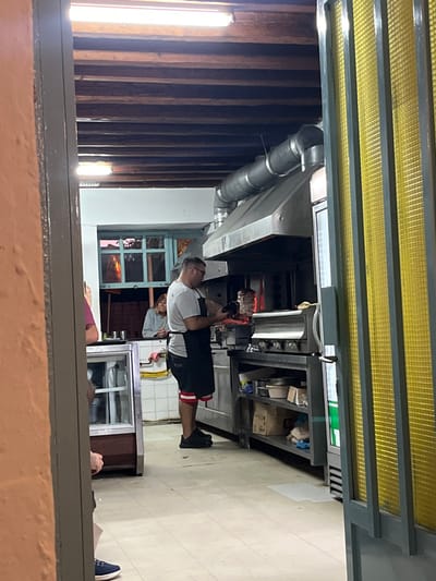 The image captures the interior of a food service establishment in Megalochori, Greece, viewed through a doorway. A male cook, wearing a grey t-shirt, black shorts with red and white stripes, glasses, and a black apron, stands centrally, operating a vertical rotisserie (gyros/souvlaki spit) from which he is carving meat. The rotisserie emits a reddish glow from heat. Above it, a large stainless steel exhaust hood is connected to an overhead metal duct system.

To the cook's left, a female with light brown hair, wearing a light-colored long-sleeved top, is visible from the chest up, positioned behind a counter against a white-tiled wall. A glass-fronted display refrigerator is located further left. On the far right, a vertical refrigerator unit with a green label is partially obscured by a yellow textured screen or door. Stainless steel counters and shelving units below the cooking station hold various containers and cardboard boxes.

The ceiling features exposed dark wooden beams with multiple fluorescent light fixtures. The floor is covered with light-colored square tiles. In the background, a light blue-framed window reveals an outdoor scene with a reddish-pink sky and dark foliage, suggesting dusk. The immediate foreground shows the lower torso and legs of a person wearing dark trousers and blue shoes, positioned within the left side of the doorway's frame, which consists of an orange-painted wall and a grey frame.