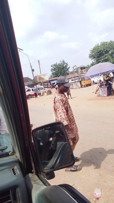 The image depicts a street scene in Igbo Ukwu, Nigeria, viewed from inside a stationary vehicle. The foreground features the red door frame and interior paneling of the vehicle, with a side mirror prominently positioned, reflecting a car seat and dashboard. The mirror's surface displays a handwritten inscription "356 KJA". A discarded plastic bottle with a pink cap lies on the unpaved ground to the right of the mirror.

The primary subject is an adult male with a dark complexion, positioned in the middle-right. He wears a black baseball cap with a light-colored logo, a short-sleeved collared shirt patterned with orange-red circular motifs on a light brown background, and dark open-toed sandals. He is looking downwards and slightly to his right, appearing to walk or stand on the dusty street.

The background shows a bustling urban environment under a cloudy sky. Multiple vehicles are present, including white mini-vans, a yellow tricycle (Keke Napep), and a motorcycle. Several individuals are visible in the distance, some standing near market stalls or goods stacked on the ground. A large blue-purple market umbrella shades an area, and a "Gemini Recharge Cards" promotional stand is visible next to a motorcycle. Utility poles with wires, a street light pole with an attached speaker, and a partially obscured sign (reading "BAPTIST CHURCH..." among other text) are also present. Buildings of varying heights and green-roofed structures line the street, interspersed with trees and sparse vegetation. Shadows indicate daylight conditions.