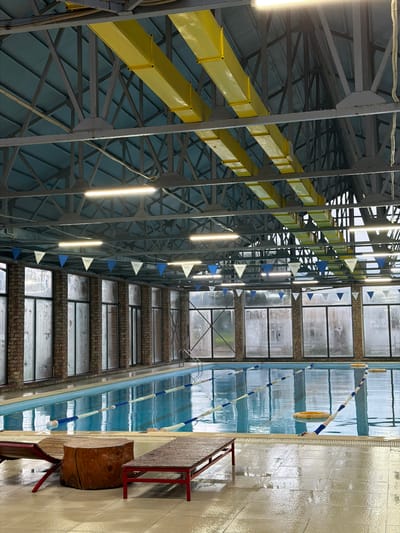 The image depicts the interior of an indoor swimming pool facility. The pool itself is filled with clear blue water and marked with lane lines. The pool area is surrounded by a tiled floor, which appears wet in places. Along the far wall, there are large windows with frosted glass, offering a view of the exterior. The ceiling is high and features exposed metal beams and yellow ductwork, along with fluorescent lighting. Triangular flags in blue and white are strung along the upper portion of the windows. In the foreground, there are two wooden benches and a large tree stump. The overall environment suggests a public or semi-public facility. The scene is set in the city of Shabdan, Kyrgyzstan.
