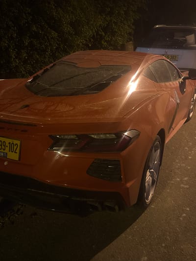 The image shows an orange Corvette parked on a street in Sdot Dan Regional Council, Israel. The license plate is partially visible. To the left, there's another vehicle and some greenery. The road surface is dark, suggesting it might be night. The car appears to be slightly dirty or wet.