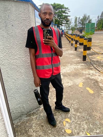 A man with dark skin, a bald head, and a beard stands in what appears to be an industrial area, possibly Kinkinau, Nigeria. He is wearing a black t-shirt, black pants, a red high-visibility vest with gray reflective stripes, and dark blue sneakers. He is holding a smartphone in his right hand and a black water bottle in his left hand. The smartphone has a case designed to look like a bookshelf. The background shows a concrete ground with yellow markings, a series of yellow and black bollards connected by chains, and some trees and buildings in the distance. The weather appears overcast.