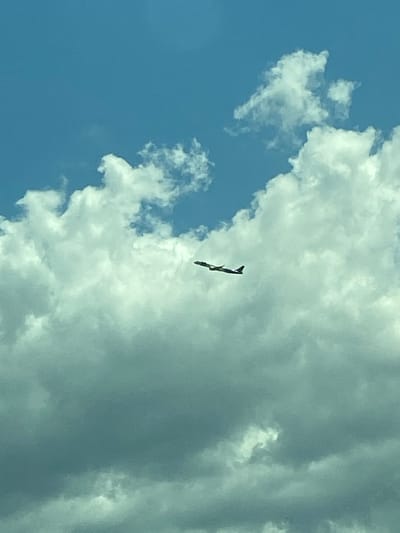 A lone airplane is captured in flight against a backdrop of a bright blue sky and scattered white clouds. The aircraft is visible in the upper center of the frame, with its wings extended. The scene does not depict any people or specific landmarks that would indicate a location within the city of Donja Cijevna, Montenegro. The focus is solely on the aircraft and the sky.