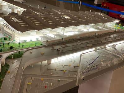 The image shows a detailed architectural model of Algiers Airport (Aeroport d'Alger), located in Dar El Beïda, Algeria. The model displays a modern airport terminal with a distinctive wavy roof structure, featuring numerous illuminated skylights. A multi-level roadway system with ramps and overpasses is visible, populated by small-scale car models in various colors (yellow, red, green, white) and miniature figures representing people. Several parking areas are marked, some with labels like "P 04". In the foreground, a clear acrylic panel with the airport's logo and name, "Algiers International", is prominently displayed. Surrounding the terminal are miniature representations of landscaping, including palm trees and green ground cover. The lighting in the model suggests an interior display environment.