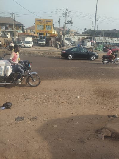 The image shows a street intersection in the city of Way, Nigeria, with a dirt road surface. There are two men on a motorcycle with bags of flour on the back. Another motorcyclist is crossing the street in the background. A black sedan is parked in the street. Several other people are walking around in the background. Behind them are buildings and a sign for "Venza Homes Real Estate." There is a lot of construction debris on the ground. The location is based on the coordinates 9.029 latitude and 7.584 longitude.