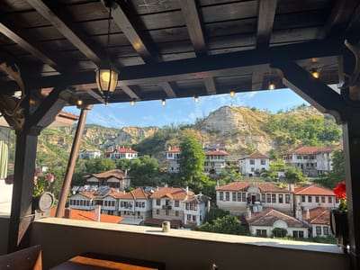 The image depicts an exterior view from a shaded balcony or veranda in Melnik, Bulgaria, overlooking the town nestled within a valley characterized by distinctive sandstone formations known as the Melnik Earth Pyramids.

In the foreground, a dark wooden ceiling structure with exposed beams and planks occupies the upper portion of the frame. A single ornate, dark-toned lantern with an illuminated bulb hangs centrally from the ceiling. A string of multiple small, illuminated Edison-style bulbs is strung across the open space beneath the main ceiling beams, framing the view. Dark wooden structural posts are visible on the left and right, with decorative carved elements. On the far left, a set of animal antlers, possibly from a deer, is mounted on a wooden base. The lower edge of the frame features a light-colored balcony railing, with a glimpse of a dark wooden table and chair below. A pot with pink flowers is visible on the left, and a small metal, cylindrical object rests on the railing. Red flowers are partially visible on the far right.

The midground showcases numerous traditional Bulgarian-style houses, predominantly white with brown trim and red-tiled roofs, built into the sloping hillsides. The architecture features multiple windows, some with balconies. Several houses have visible solar panels on their roofs. Green trees and dense vegetation are interspersed throughout the residential area, obscuring some parts of the buildings. A sign "ART & WINE SHOP" is discernable on one building's facade.

In the background, the unique, eroded sandstone formations of the Melnik Earth Pyramids rise prominently, covered in patches of green foliage, under a clear blue sky with sparse white clouds. A small, cross-like structure or beacon is visible on the peak of one distant pyramid. No people are visible in the scene.