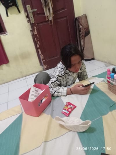 A young person with dark hair is lying on the floor, looking at a smartphone. They are wearing a long-sleeved shirt with a black and white checkered pattern. The setting appears to be indoors, with tiled flooring and a wooden door visible in the background. A rolled-up rug is leaning against the wall.

On the floor around the person are several objects. A pink rectangular basket holds some items, including what appears to be packaging with text. Next to the basket is a red packet of candy, with the word "BISKIN" visible. A white, woven hat lies on the floor as well. To the right, a wicker basket contains several small bottles, some of which are spray bottles with colored caps, suggesting toiletries or cosmetics.

The lighting in the image is dim, suggesting it might be evening. The overall scene is casual and domestic. A timestamp in the lower right corner indicates the date "26/06/2026" and the time "17:50". The location context is Sukaraja, Indonesia.
