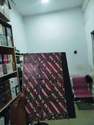 A person's hand holds a hardcover book with a distinctive red and white striped pattern and a dark abstract design. The book is positioned in the foreground, obscuring the lower portion of a shelf filled with various items, including what appear to be boxes and cylindrical containers, some with gold accents. To the right of the book, a blurry red chair is visible. The environment suggests an indoor setting, possibly a shop or storage area in Uselu, Nigeria, with shelves lining the walls. The lighting is somewhat dim, originating from above.
