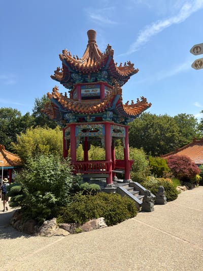 The image depicts a vibrant, ornate Chinese-style pavilion set within a lush garden. The pavilion features a multi-tiered, curved roof with intricate detailing in red, blue, and gold, adorned with dragon motifs and decorative finials. Supporting red pillars frame open sections of the structure, revealing painted panels. To the right of the pavilion, a stone staircase with carved lion or dragon heads leads upwards, bordered by neatly trimmed green bushes and rugged rocks. A textured, light-colored pathway runs alongside the garden, with a subtle brick border.

Behind the pavilion, a variety of trees and shrubs create a dense, green backdrop, with a hint of red foliage on the right. The sky is a clear, bright blue with a few wispy white clouds, suggesting it is daytime and the weather is pleasant. Two neutral-colored banners with circular emblems are visible in the upper left of the frame, adding to the cultural context. In the lower right portion of the image, two people can be seen walking on the pathway, one wearing a straw hat. The overall atmosphere is serene and picturesque, likely a tourist attraction or a cultural garden. The scene appears to be set in Saint-Aignan, France, given the provided location context. No visible text other than the emblems on the banners.