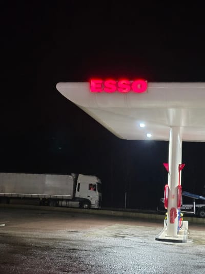 The image shows an Esso gas station at night. The red "ESSO" sign is illuminated on the station's canopy. A white semi-truck is parked at the gas station, and a wet concrete surface is visible. The lighting is generally dark, with the bright gas station sign providing the main source of illumination.