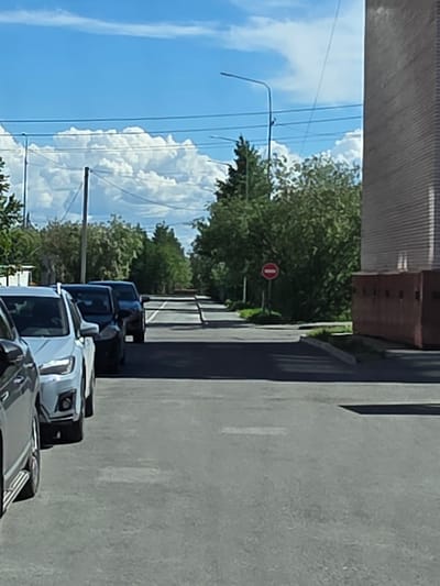 A street scene in Salekhard, Russia, features a line of cars parked on the right side of a paved road. The road extends into the distance, lined with trees on both sides. In the background, a large building with a textured facade is visible on the right. A "no entry" sign is posted on the left side of the road. The sky is blue with scattered white clouds.