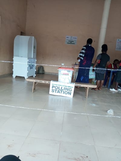 This image depicts an indoor polling station located in the city of Ajalli, Nigeria. The environment features light brown, textured walls with minor cracks and a light-colored tiled floor. A white rope defines a boundary across the foreground and midground.
On the left, a white, partially enclosed polling booth displays "POLLING ZONE" and a green logo, likely from the Independent National Electoral Commission (INEC). A dark-colored backpack rests on the floor behind it. In the midground, a simple wooden bench, partially inscribed with "LEGION OF M" and "URHU," supports a clear plastic ballot box with a red lid, labeled "BALLOT BOX" and "INEC," containing documents. A white sign with black text reading "INDEPENDENT NATIONAL ELECTORAL COMMISSION POLLING STATION" is positioned on the floor in front of the bench.
Three individuals are present on the right. Two women stand with their backs to the camera; one wears a long dark blue dress with white patterns, and the other has a dark patterned top and dark skirt. A third person, wearing a red top, is partially visible, seated at a blue table on the far right.
A poster on the wall states "INEC FC 20 C VOTING IN PROGRESS." Two white electrical outlets are visible on the wall, and a white vertical pillar extends upwards on the right side. Small pieces of white debris are scattered on the tiled floor.