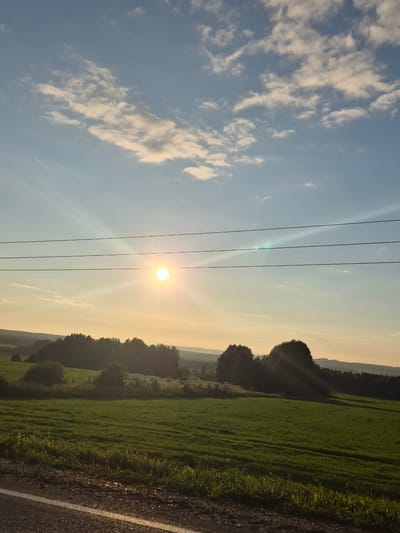 This is a vertically oriented, wide-angle photograph taken at sunset in Orekhovaya Gora, Russia. The scene depicts a rural landscape with a prominent power line running horizontally across the frame, dividing the sky and the ground.

The sky is a gradient of blue on the left, transitioning to a soft yellow and orange hue near the horizon, indicating the setting sun. Wispy white clouds are scattered across the blue portion of the sky. The sun itself is a bright, radiant orb, casting a warm glow and visible sunbeams.

The lower half of the image features a vibrant green field, possibly a pasture or farmland, stretching into the distance. A line of dark trees forms a silhouette against the lighter sky along the left edge of the field. To the right of the field, a dirt road or path with a white line marking is visible, suggesting the edge of cultivated land. The overall mood is peaceful and serene, characteristic of a rural sunset.

There is no visible text in the image.