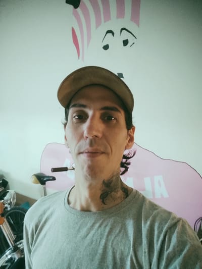 A man wearing a tan baseball cap and a gray t-shirt is posing for a selfie. He has a tattoo on his neck. Behind him is a motorcycle and a mural with pink and white stripes and black oval shapes on a white background. The mural also appears to have pink shapes with white letters. The location is Remedios de Escalada, Argentina.