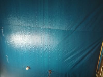 The image shows a blue tarp covering what appears to be a structure or object. The tarp has a repeated "SAKURA" logo with a floral design in the lower left and upper left corners. There are dark linear patterns, possibly seams or folds, running vertically. A light bulb is attached on the lower left of the tarp. In the bottom center of the tarp, there appears to be another attachment point. The environment is dark. The exact location in Kruengmanyang, Indonesia, cannot be determined from the visual information provided.