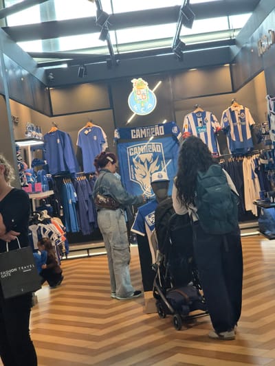 The image captures the interior of a store, likely a sports merchandise shop, located in Moreira, Portugal. Various individuals are present, including customers browsing through the items on display. The main focus is on apparel, specifically soccer jerseys, displayed along the wall on hangers. The color scheme of the merchandise is predominantly blue and white.
One individual is pushing a stroller, and another appears to be examining a product with "FASHION TRAVEL GATE" imprinted on it. The environment is well-lit, with natural light streaming in from a window on the left. The flooring is a light-colored wood.
The prominent logo, depicting the emblem of FC Porto, is displayed on the wall, indicating a sports store that sells FC Porto merchandise.