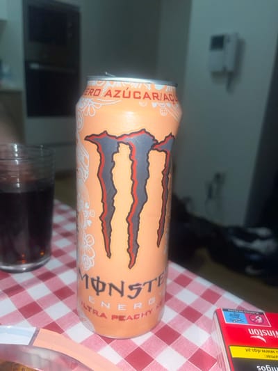 This image depicts a close-up of a peach-colored "Monster Energy Ultra Peachy Keen" beverage can, positioned on a red and white checkered tablecloth. The can features a prominent black, grey, and orange Monster logo, surrounded by white decorative patterns including peace signs and flowers. The text "ZERO AZÚCAR/AÇÚCAR" is visible near the top, and condensation is present on the can's surface. To the immediate left, a partially obscured glass filled with a dark liquid is visible. To the right of the can, a red pack of "Winston" cigarettes, partially inverted, rests on the tablecloth. The background is dimly lit and blurred, revealing a dark rectangular object on the left, a light-colored wall, and a white wall-mounted device, possibly a thermostat or intercom, on the right. Indistinct dark items are present on a lower surface in the far right background. No individuals are present in the scene, which is located in the city of Pedrouços, Portugal.
