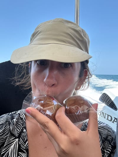 The image displays an adult individual situated on a motorized boat in Pak Nam, Thailand. The person, positioned from the chest up, wears a tan wide-brimmed hat and a black and white patterned top. Their dark eyes are wide open, directed towards the viewer. The individual holds two individually plastic-wrapped muffins in their left hand, with their mouth pressed against the clear plastic of the muffin on the left. A small, light-colored earring is visible in their right ear. The background features a clear blue sky meeting a body of blue water, with a white wake trailing behind, indicating the boat is actively moving. A silver metal pole of the boat structure rises on the right side of the frame, alongside a white boat panel featuring a black grille and the partially visible brand name "HONDA."