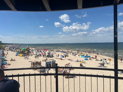 The image captures a bustling beach scene in Międzyzdroje, Poland, on a sunny day. The foreground is dominated by a black metal railing, through which a wide expanse of golden sand and the Baltic Sea is visible. A person in a dark blue shirt is seated, facing away from the camera, looking out at the beach.
The beach itself is densely populated with people enjoying various activities. Numerous beach umbrellas, tents, and chairs are scattered across the sand, providing shade for sunbathers. A colorful children's playground, complete with slides and climbing structures, is a prominent feature in the mid-ground, with children actively playing. People are seen walking, lounging, and swimming in the calm, blue waters of the sea. The coastline stretches into the distance, lined with trees and buildings in the far background, suggesting a developed resort area. The sky is a bright blue, dotted with fluffy white clouds. The overall atmosphere is one of a popular summer vacation destination, filled with leisure and recreational activities.