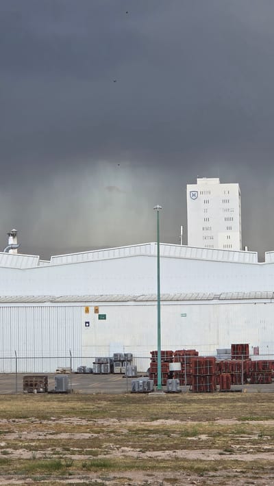 The image displays an industrial area in La Tijera, Mexico, under a dark, overcast sky. In the mid-ground, a large, white corrugated metal factory building is prominent, featuring multiple warning signs and green signage on its facade. To the right and behind this structure, a taller, rectangular white building with a blue 'H'-like logo on its upper left side is visible, characterized by numerous vertical architectural details. A single green lamppost stands in the lower mid-ground, positioned in front of a chain-link fence. Behind the fence, on a paved surface, are stacks of industrial materials: gray block-like components are arranged on pallets to the left, and numerous reddish-brown cage-like structures are stacked on pallets to the right. The foreground consists of dry, patchy earth with sparse vegetation. No individuals or active processes are depicted.