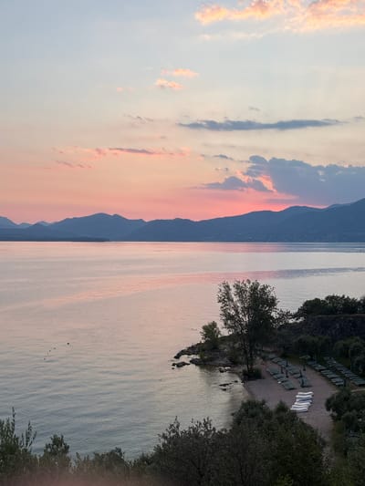 This image depicts a serene sunset scene over Lake Garda, Italy. The sky is painted with soft hues of pink, orange, and blue, with scattered clouds catching the last light of the day. In the foreground, a small beach is visible with rows of empty lounge chairs and several white kayaks neatly arranged on the sand. Lush green trees and foliage line the edge of the beach, and rocky outcrops break the shoreline. The calm water of Lake Garda stretches towards a distant mountain range, silhouetted against the colorful sky. The overall impression is one of tranquility and natural beauty.