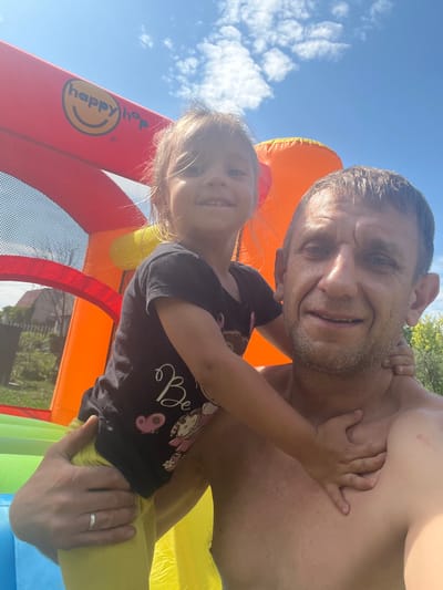 A selfie taken outdoors on a sunny day shows a man and a young girl, likely father and daughter, with a bouncy castle in the background. The girl, in a black t-shirt with a graphic design, is positioned in front of the man, with her arms around his neck and chest. The man, shirtless, is smiling at the camera. The bouncy castle is red and orange, with a "happy hop" logo visible. In the background, a house and trees suggest a residential area. The image appears to have been taken in Koniukhi, Russia, a small village.