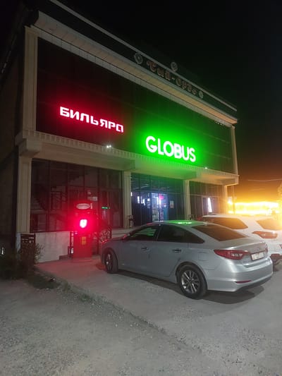 A modern, multi-story building at night, likely a commercial establishment, is illuminated with neon signage. To the left, in bright red neon, is the Cyrillic word "БИЛЬЯРД" which translates to "Billiard". To the right, in bright green neon, is the word "GLOBUS". The building has large glass windows on the ground floor. In front of the building, a red, upright machine with a lit red display is visible. A silver sedan is parked in front of the building, facing away from the camera, with its rear visible. To the right of the sedan, the rear of a white SUV is partially visible. The ground in front of the building is rough and unpaved, with some vegetation along the left edge. The sky is dark, indicating nighttime. The building's architecture appears contemporary, with clean lines and decorative elements along the roofline. The overall impression is that of a well-lit commercial area at night.