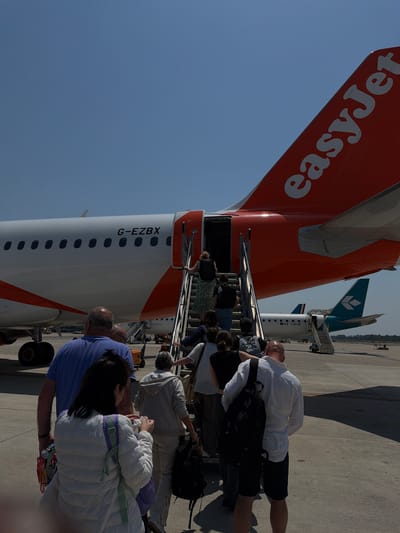 A group of people are boarding an EasyJet airplane via stairs on a sunny day at an airport. The airplane is white with orange accents and has the registration "G-EZBX" visible on its side. Another smaller airplane with a green tail and "I-ADJN" on its side is parked behind it. The scene is taking place on the tarmac, with a clear blue sky overhead. The people are dressed in casual attire, with some carrying luggage. The airport appears to be in Peschiera Borromeo, Italy.