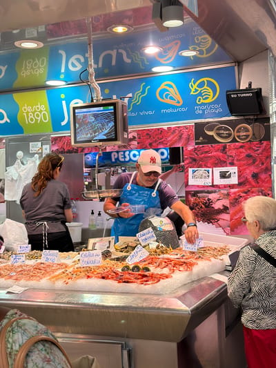 This is a street-level, medium shot of a seafood stall at a market in Alboraia, Spain. The stall is filled with various types of seafood displayed on ice. A man in a blue apron and a white baseball cap is serving a customer, an older woman wearing a black and white striped shirt and red pants. The man is holding a plastic container of seafood. Signs with prices are displayed on the ice. In the background, there are promotional signs with images of seafood and text in Spanish, indicating "mariscos de playa" (beach seafood). A digital screen displays prices and images of food. The stall is brightly lit, and the overall atmosphere is that of a bustling market.