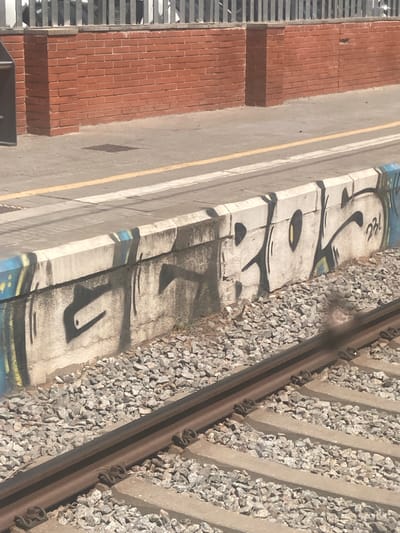 The image shows a section of a train platform in Cerdanyola del Vallès, Spain. A concrete wall at the edge of the platform is covered in graffiti. The graffiti is done in black and white with some blue and yellow accents. The wall is located adjacent to a set of train tracks covered in gravel. In the background, a red brick wall and a metal fence are visible. The scene appears to be captured in daylight.