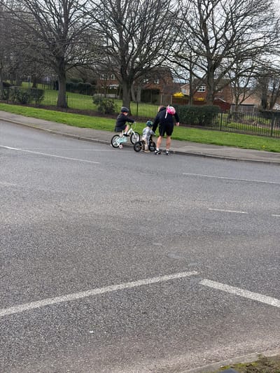 The scene is located in the city of Rotherham, United Kingdom. Three individuals are positioned on a paved footpath adjacent to an asphalt road. An adult male, wearing a black shirt, black shorts, black sneakers with white accents, and a pink backpack, is observed standing next to a young child. This child wears a green helmet and a plaid shirt, and is seated on a small orange-brown tricycle. A second child, wearing a black helmet and a dark jacket, is riding a light blue bicycle equipped with white training wheels, positioned slightly ahead and to the left of the adult and younger child.
The asphalt road in the foreground displays white painted markings, including a dashed line and a solid line forming a junction or 'give way' indication. Adjacent to the footpath is a green grass verge with scattered yellow flora, bordered by a black metal fence. In the background, numerous large, bare deciduous trees are visible, indicating a non-summer season. Beyond the trees, red brick residential houses with visible windows are present. The sky above is overcast, suggesting a cloudy daytime.
