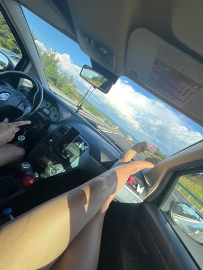 The image depicts the interior of a car during daytime, with a focus on the dashboard and the legs of passengers. The sun is shining brightly, casting shadows on the dashboard and the legs. The car appears to be moving on a highway, as indicated by the road visible through the windshield and the side windows. The dashboard is visible, showing the steering wheel with a "FIAT" logo, gear shifter, and various control buttons. A smartphone is mounted on the dashboard. The legs of two individuals are visible, one resting on the dashboard and the other extending towards the camera. The scene suggests a road trip or travel, with the environment outside the car indicating a rural or roadside setting with trees and a cloudy sky. The text on the sun visor, "BALIOVCI, BULGARIA," indicates the location.