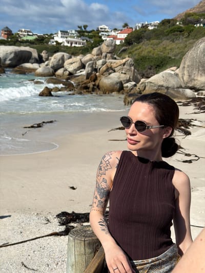 The image features a young woman looking out over a coastal landscape, captured likely in Simon's Town, South Africa. She has dark hair tied back and is wearing round, dark sunglasses, a dark brown ribbed sleeveless top, and a light-colored patterned skirt or sarong. Her left arm is extensively covered in black tattoos with geometric and floral designs. She is leaning on a wooden railing, positioned on the right side of the frame, with her face gently turned to her left, displaying a slight smile.

The scene behind her unfolds into a bright, sunny daytime beach environment. A sandy shore with gentle waves and scattered dark seaweed stretches from the foreground towards the left. The coastline is marked by numerous large, smooth grey boulders. Beyond the rocks, a verdant green hillside rises, dotted with white houses featuring reddish-brown roofs, indicating a residential area built into the landscape. The sky above is a clear blue with some soft white clouds. The overall impression is one of calm and relaxation by the sea. No visible text is present in the image.