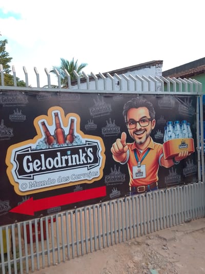 A banner advertising "Gelodrinks - O Mundo das Cervejas" is displayed on a metal fence in Cáceres, Brazil. The banner features a cartoon character of a man with glasses, a beard, and an orange shirt, holding a six-pack of bottled beverages and giving a thumbs-up. To the left, there's a graphic of three beer bottles on ice, with the text "Gelodrinks" prominently displayed. A large red arrow points to the left, indicating direction. The background of the banner is black, with repeated logos and the slogan "Gelodrinks - O Mundo das Cervejas" in a repeating pattern. The fence is made of vertical grey metal bars. In the background, there's a building with a tiled roof and some vegetation, including a palm tree. The sky is visible above the banner.