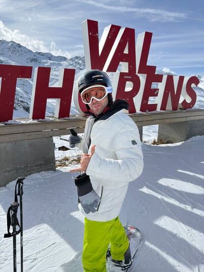 A male individual stands on a snowy slope in Val Thorens, Les Belleville, France, posing in front of a large, red and white block letter sign spelling "VAL THORENS." He is wearing a black ski helmet with orange goggles, a white quilted ski jacket over a black hoodie, bright neon green ski pants, and grey gloves. His left hand is raised, forming a "shaka" gesture. Two black ski poles are planted in the snow in the lower left foreground. The individual is positioned with a snowboard partially visible at his feet on the right. The background features expansive snow-covered mountains under a clear blue sky with scattered white clouds. The ground immediately around the sign's base shows some patches of dry vegetation not covered by snow.