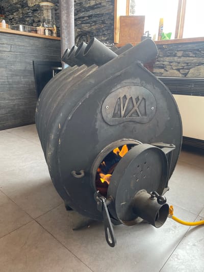 The scene depicts an interior space within a structure located in Juta, Georgia. A large, barrel-shaped, black metal wood-burning stove is centrally positioned on a light gray tiled floor. The stove's front loading door is partially open, revealing active orange and yellow flames from burning material inside. A black metal latch mechanism is attached to the door. A yellow flexible hose is connected to a fitting on the lower right of the stove's front assembly. A custom metal "MAX" logo, secured by two screws, is affixed to the stove's right side. A large vertical stovepipe extends upward from the rear-center of the unit, and four capped pipe outlets are visible along the top-rear. The background features a dark, textured stone wall. To the left, a dark wood-paneled counter displays several clear glass jars and bottles. A window, admitting natural light, is visible on the right side of the frame, with a white horizontal radiator positioned beneath it against the stone wall. No individuals are present in the image.